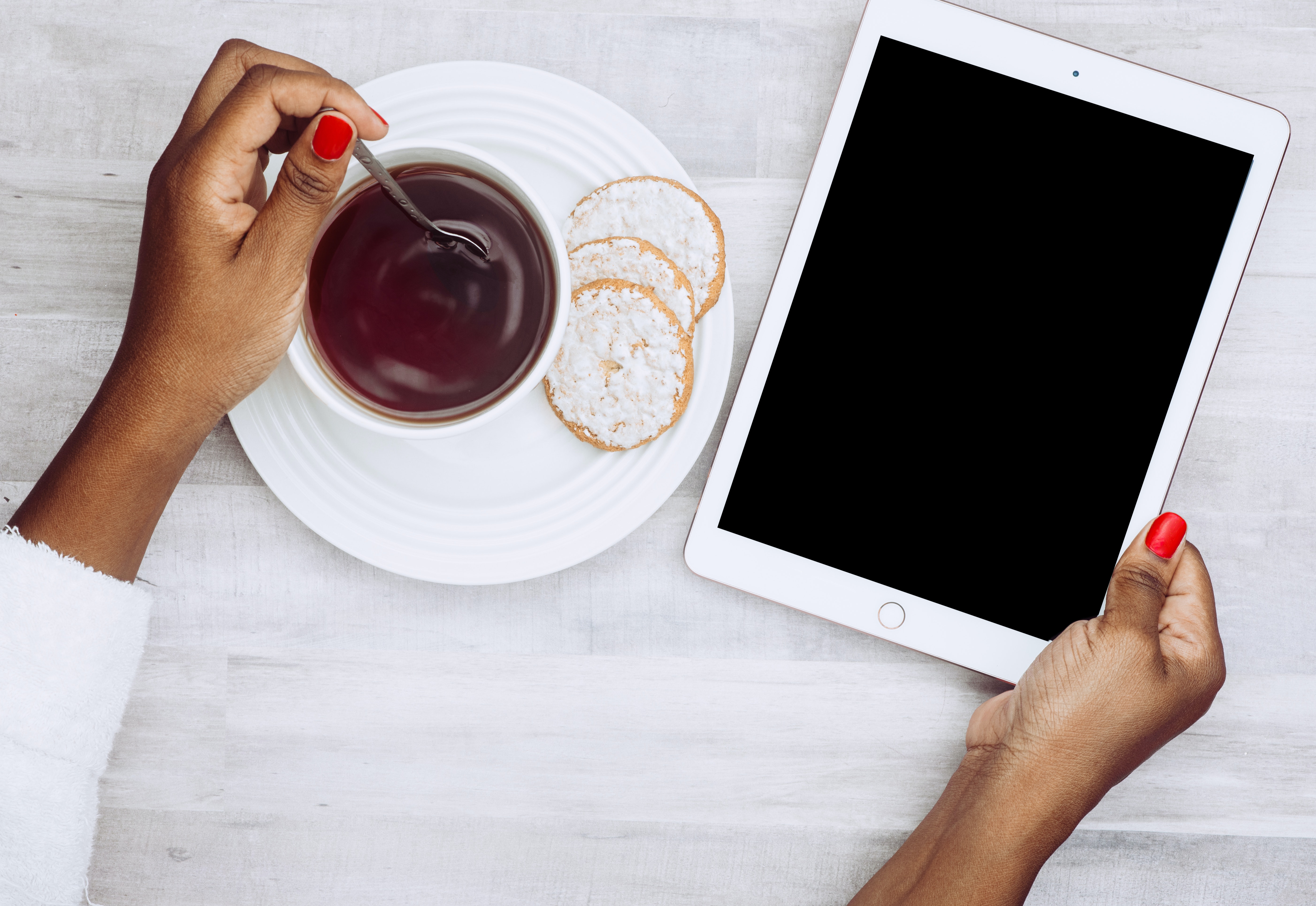 En kaffekopp och ett fat med tre kakor, en hand som rör med en sked i kaffekoppen och en hand som håller i en iPad.