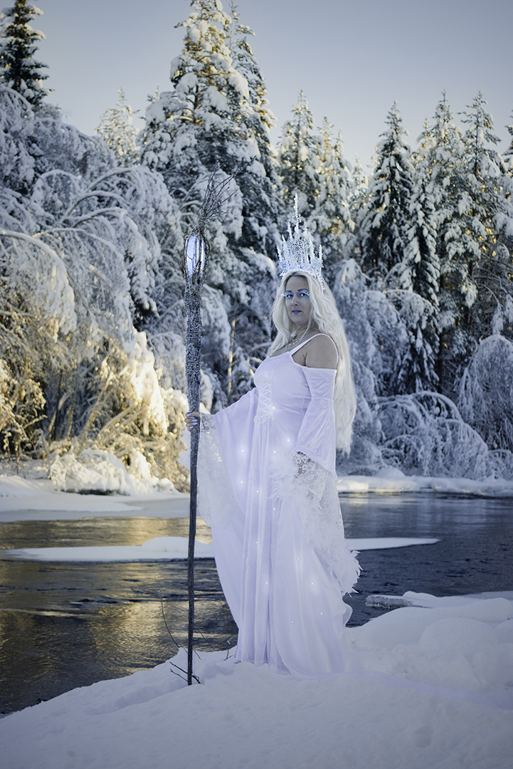 Isdrottningen. Kvinna med långt blond hår klädd i en skimrande vit klänning står intill en älv i ett snötäckt vinterlandskap. Hon har en iskrona på huvudet och håller i en stav med magisk eld i toppen som är längre än hon själv är.