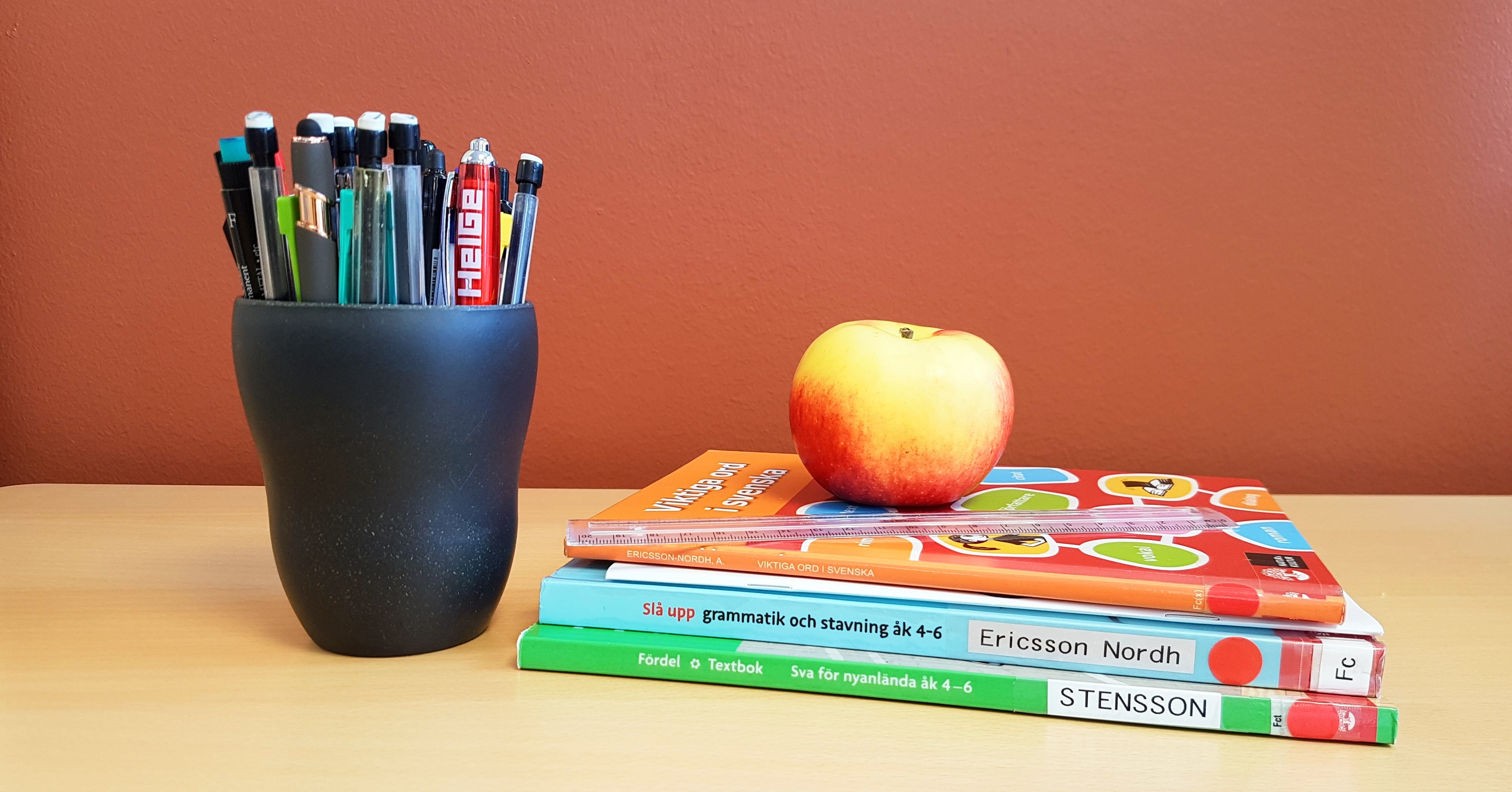 En trave böcker, med en linjal och ett äpple ovanpå böckerna, bredvid en burk med pennor.