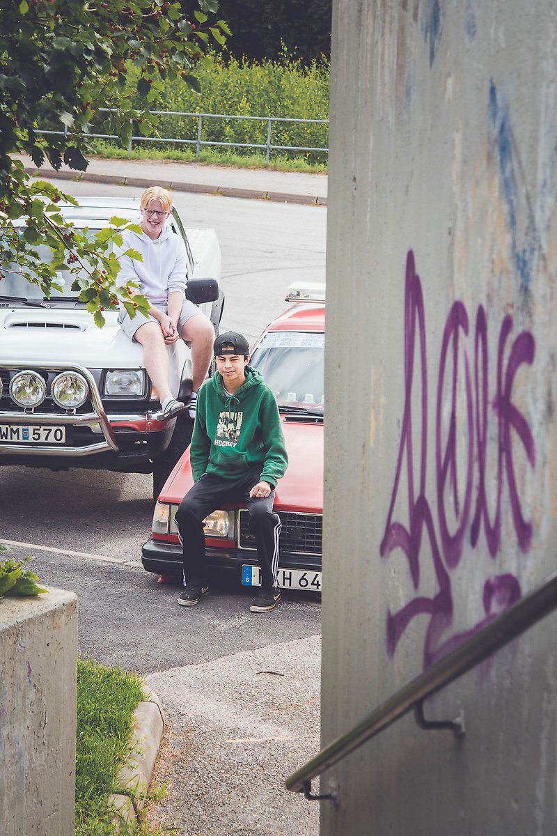 Ture och Albin sitter på huvarna på sina EPA-traktorer på en asfaltsparkering. På en vägg i förgrunden är det målat graffiti.