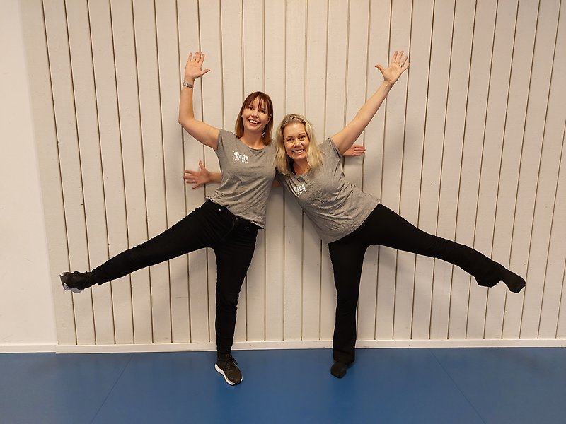Två kvinnor i grå t-shirt och svarta byxor står framför en vit vägg och sträcker ut ben och armar på ett lekfullt sätt. 