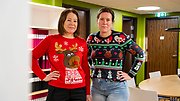 Två kvinnor i kontorsmiljö iförda jultröjor. Den ena tröjan är röd med ett renhuvud och snöflingor på. Den andra är mörkblå med mönster av granar, renar och snögubbar.