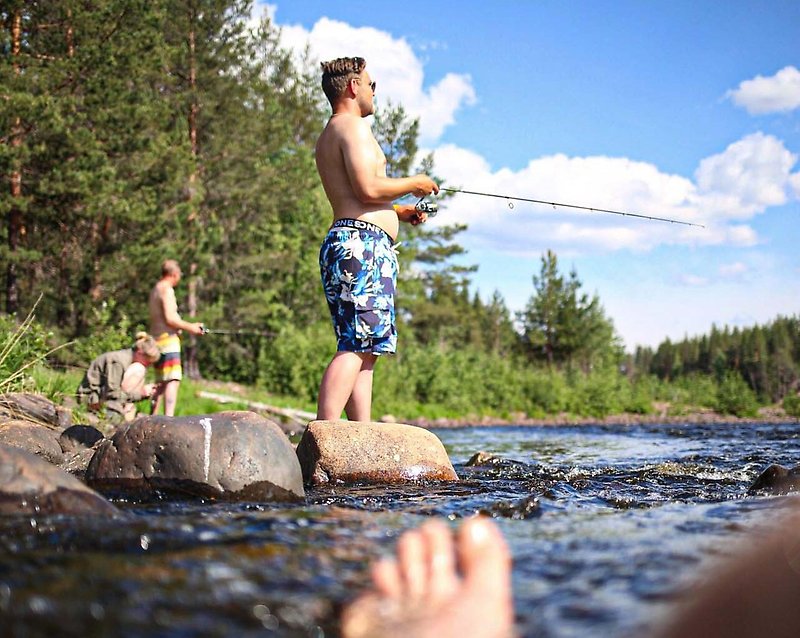 Ett gäng ungdomar står vid ett vattendrag och fiskar under blå himmel.