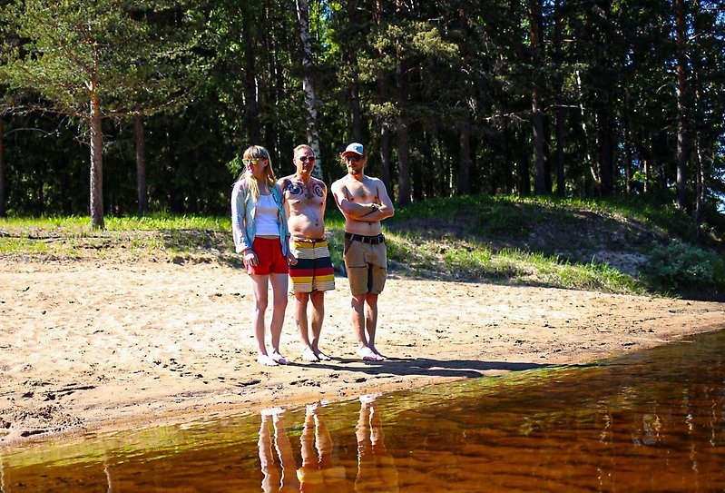 Tre ungdomar står på en strand vid vattenbrynet med en skog bakom sig.