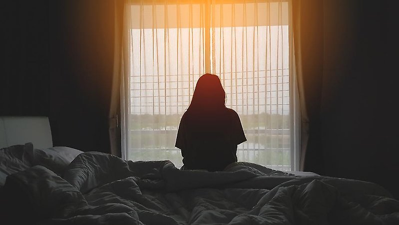 En kvinna som sitter i en säng och tittar ut genom ett fönster i motljus