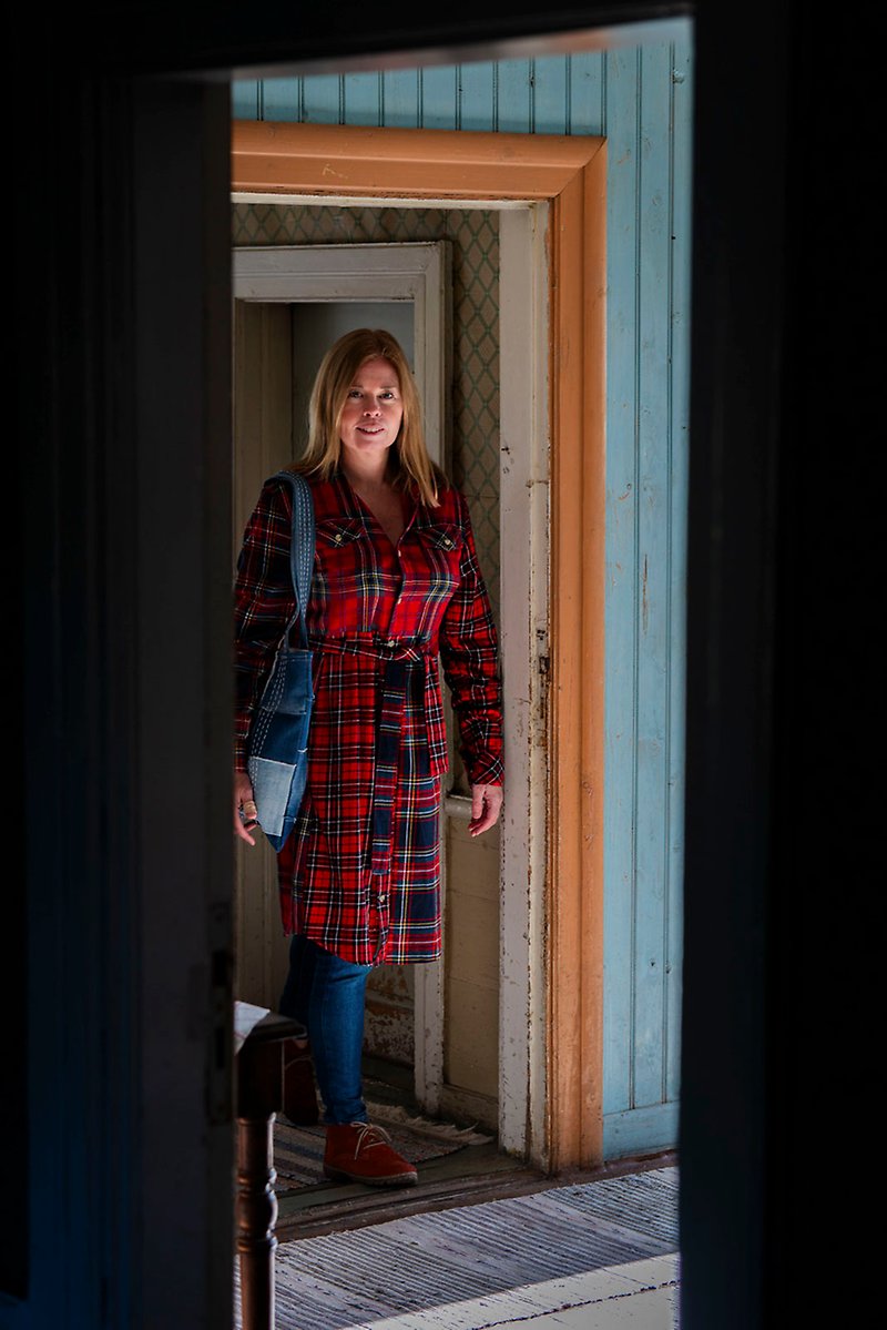 Malin Svedberg står i en dörröppning mellan två rum i ett gammalt hus iförd en röd-svart-rutigt rock och en väska gjord av jeanstyg som hänger på hennes axel.