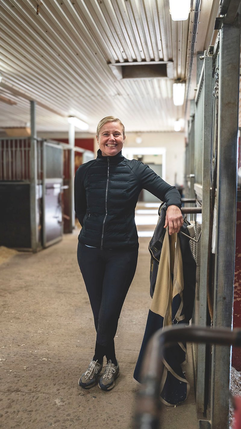 Maria Eriksson står och lutar sig mot väggen i ett stall och ler mot kameran.
