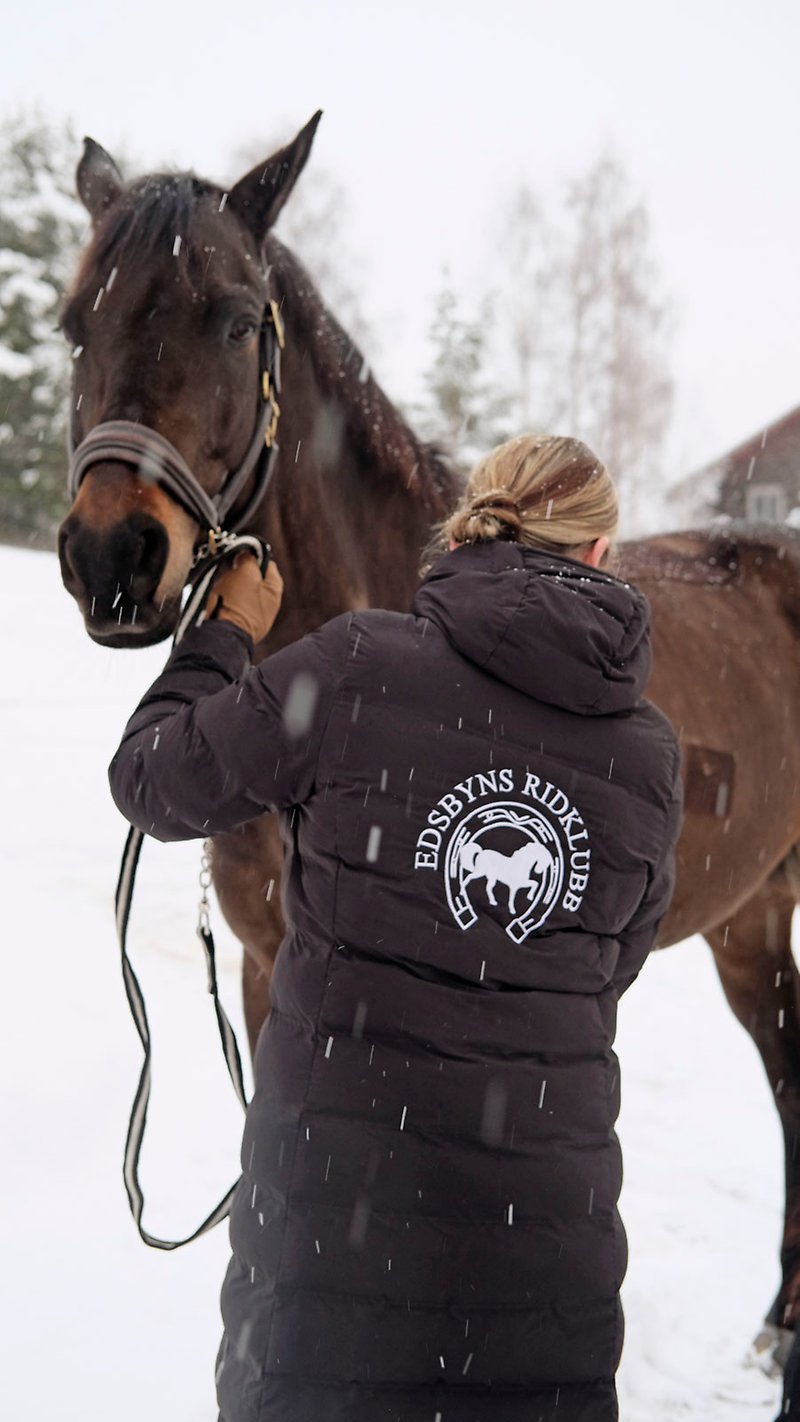 Maria leder sin mörkbruna häst på en snötäckt gård. Hennes rygg är vänd mot kameran. På ryggen på hennes mörka vinterjacka sitter en vit logotyp för Edsbyns Ridklubb.