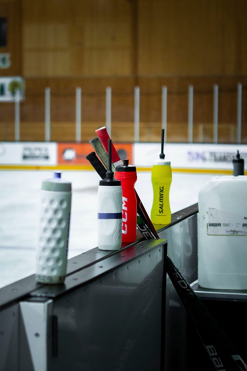 Vattenflaskor i olika färger står på sargen vid en inomhushockeyplan. Mot sargen står några hockeyklubbor lutade.