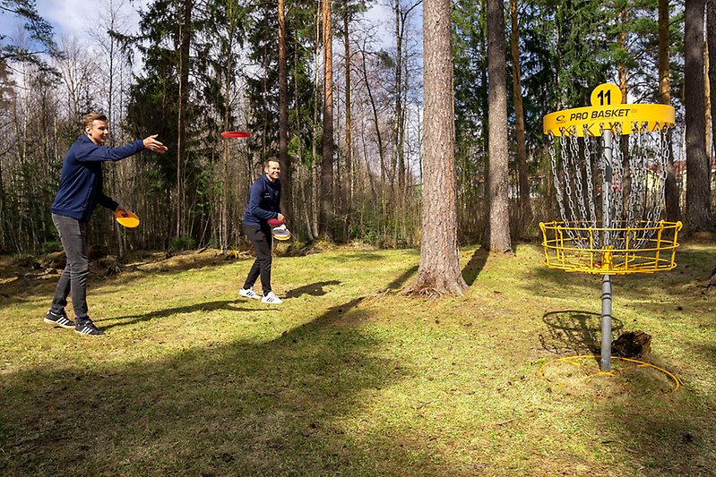 Felix Karsbo och Calle Kopparmalms står i skogen och kastar discar mot en discgolfkorg.