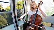 Kristina Winiarski sitter och spelar cello i en linbanevang ovanför ett skogslandskap.