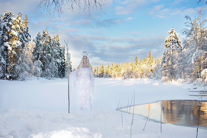 Isdrottningen. Kvinna med långt blond hår klädd i en skimrande vit klänning står på en frusen sjö i ett snötäckt vinterlandskap. Hon har en iskrona på huvudet och håller i en stav med magisk eld i toppen som är längre än hon själv är.