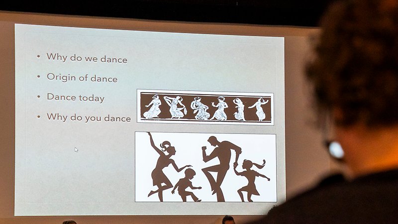 Powerpointpresentation på en bioduk. I presentationen är en punktlista med punkterna "Why do we dance", "Origin of dance", "Dance today", och "Why do you dance". Bredvid punktlistan är några illustrationer av människor som dansar. I förgrunden syns baksidan av en persons nacke.