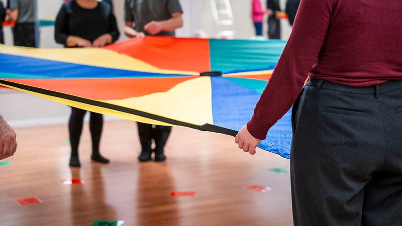 Personer står runt en stor flerfärgad duk och håller upp den i luften. Färgade lappar med siffror på är utspridda på golvet.