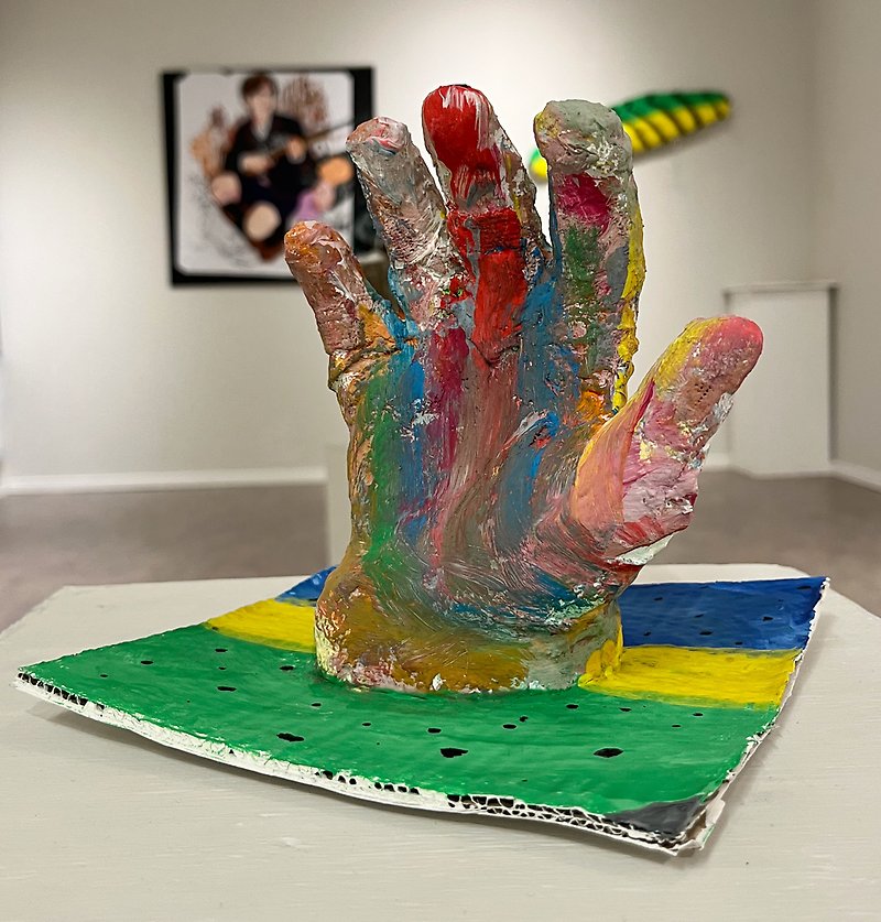 Ett färgglatt, tredimensionellt model av en hand i gips på en grund av kartong. Grunden år målat randig i blå gul och grönt med svarta prickar. Handen är målat i alla möjliga färger blandat ihop utan något mönster.