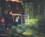 Christian sitter längst bak i sin van ute i skogen tillsammans med sin hund och båda blickar ut över skogsvägen.