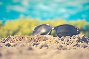 I sanden ligger ett par solglasögon och träbrickor med bokstäver på som tillsammans bildar ordet "summer".