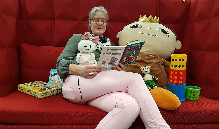 En kvinna sitter i en soffa och läser en barnbok, omgiven av sagofigurer och spel.