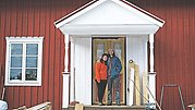 Will och Charlotte står på verandan till sin gård, omgiven av plankor, bygglampor och verktyg.