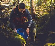 Joakim Larsson sitter på huk i ett vattendrag i skogen och skopar upp vatten i en plastskål.