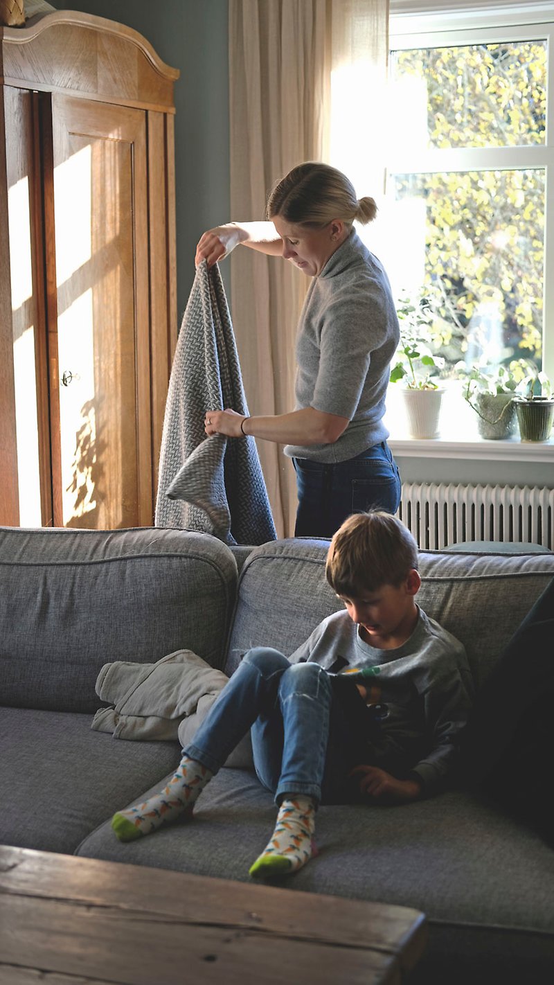 Elin viker ett tyg i vardagsrummet medan hennes son sitter i soffan med en surfplatta.